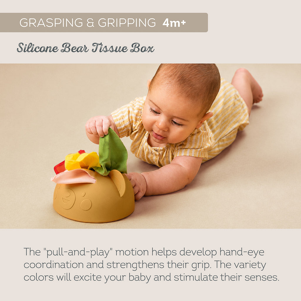 Baby lying on floor pulling fabric tissue from Taf Toys soft silicone bear tissue box for 4M+ fine motor grasping
