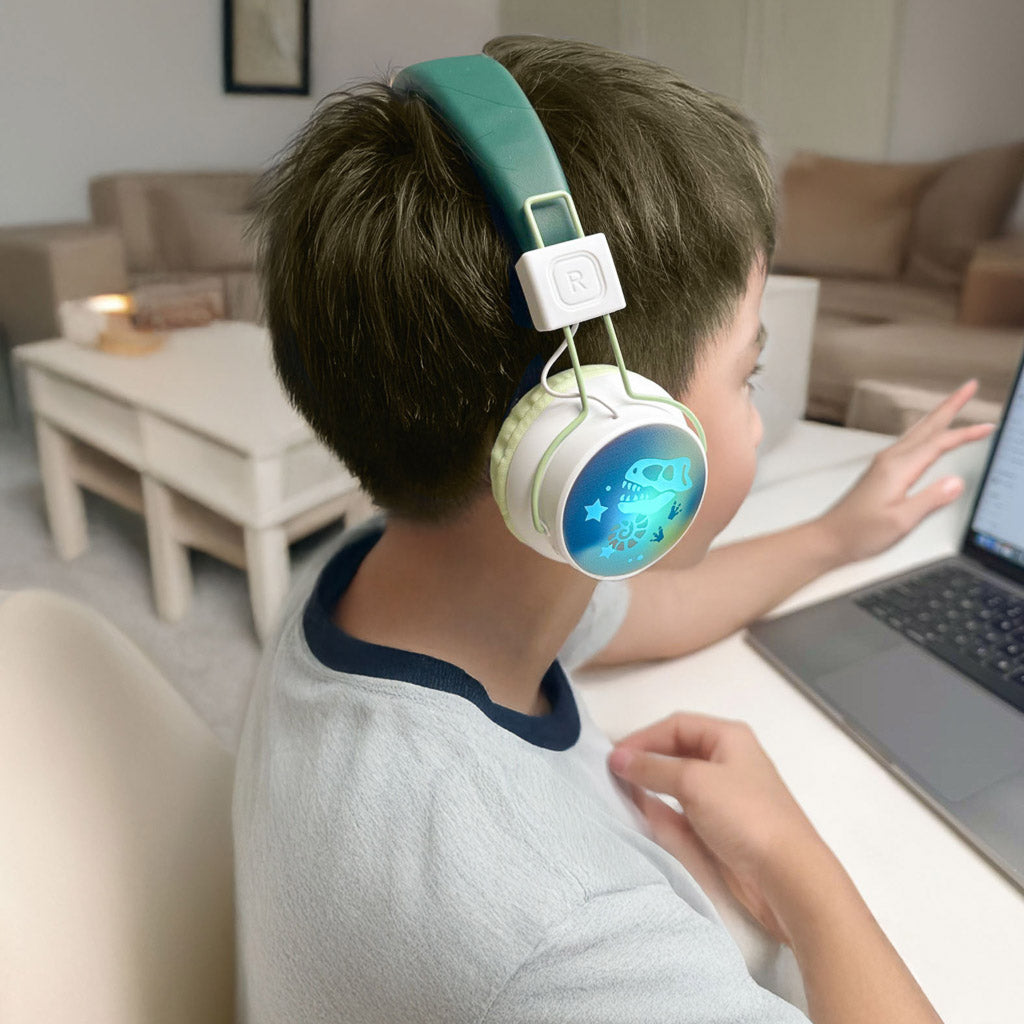 A child wearing a pair of Marckids ProtectedSound 85dB safe volume wireless Bluetooth kids headset, seated at a desk using a laptop in a bright living room.